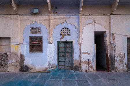 Pushkar, India - March 18, 2022: The weathered exterior of an old house features faded paint, a central arched doorway with a wooden door, two wooden windows, and visible cracks and peeling plaster.のeditorial素材