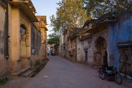 Bundi, India - March 19, 2022: A narrow street lined with weathered historic buildings featuring ornate arches and faded paint, with a tricycle parked in the foreground and trees visible in the background during daylight.のeditorial素材