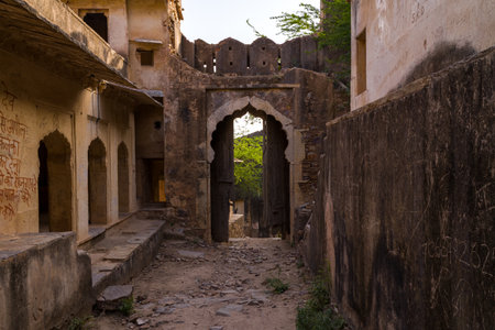 Taragarh Fort, India - March 20, 2022: The image shows a narrow stone alleyway with uneven ground and weathered plastered buildings, leading to an arched wooden gate set in a stone wall, with Hindi writing visible on the left wall and greenery beyond the のeditorial素材