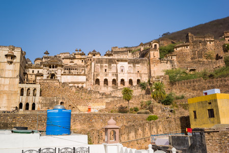 Garh Palace, India - March 20, 2022: The historic Garh Palace complex is seen with its multi-story stone and plaster structures, arched windows, and defensive walls on a hillside, with a blue water tank and yellow building in the foreground under a clear のeditorial素材