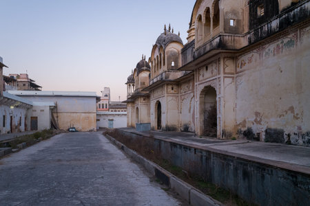 Pushkar, India - March 18, 2022: An empty paved street runs alongside weathered temple buildings with ornate arches, domed rooftops, faded wall paintings, and peeling paint at Sri Rangnath Swamy Temple during dusk.のeditorial素材