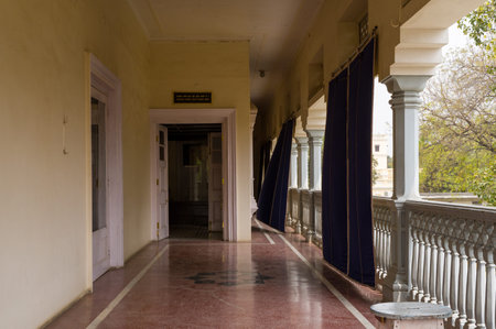 Prayagraj, India - March 21, 2024: The image shows an interior corridor of Anand Bhawan Museum with red flooring, white columns, a balcony railing, blue curtains, a small white stool, and an open doorway during daylight.のeditorial素材