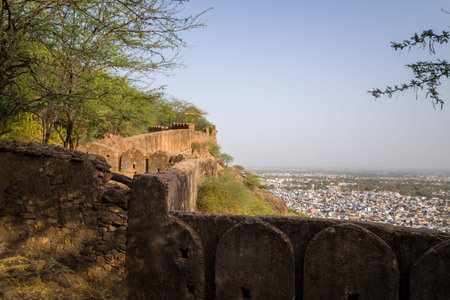Bundi, India - March 20, 2022: The image shows a weathered stone fortification wall with arched crenellations at Taragarh Fort, with trees and vegetation in the foreground and a wide view of Bundi city in the background under clear daylight.のeditorial素材