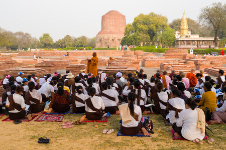 Sarnath, India - March 19, 2024: A large group of people sit on mats in front of a Buddhist monk leading a prayer or teaching session among exposed archaeological brick remains, with the Dhamekh Stupa and Mulagandha Kuti Vihar temple visible in the backgrのeditorial素材