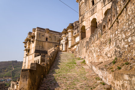 Bundi, India - March 20, 2022: The image shows a steep cobblestone access path leading uphill between weathered stone and plaster walls of Garh Palace, with arched windows, battlements, and hillside vegetation visible under a clear sky.のeditorial素材