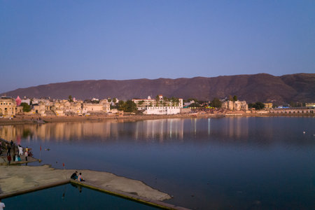 Pushkar, India - March 18, 2022: The ghats and historic buildings of Pushkar are reflected in the calm waters of Pushkar Lake at dusk, with people visible along the lakeshore and hills in the background.のeditorial素材