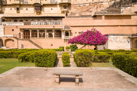 Bundi, India - March 20, 2022: The image shows a marble bench in the foreground, a manicured garden with trimmed hedges, and a flowering tree with bright pink blossoms in front of the historic exterior of Garh Palace under a clear sky.のeditorial素材