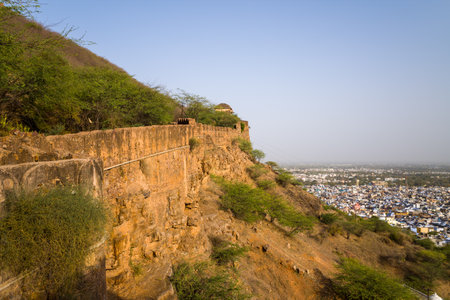 Bundi, India - March 20, 2022: The image shows a weathered stone fortification wall running along a steep hillside at Taragarh Fort, with green vegetation on the slope and a wide view of Bundi city in the background under clear daylight.のeditorial素材