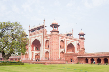 Agra, India - March 11, 2024: The image shows a side view of the main entrance gate of the Taj Mahal complex, featuring red sandstone architecture with white marble inlay, arched entrances, decorative towers, and a large tree on the lawn in the foregroundのeditorial素材