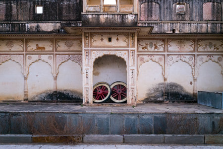 Pushkar, India - March 18, 2022: The exterior wall of Sri Rangnath Swamy Temple features ornate painted arches, faded animal motifs, and two large wooden cart wheels placed under a central archway.のeditorial素材