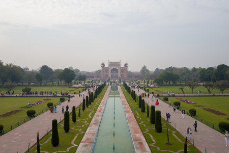 Agra, India - March 11, 2024: The main red sandstone entrance gate of the Taj Mahal complex is seen in the distance, reflected in the central pool, with rows of cypress trees and groups of people walking along the Mughal garden pathways under a cloudy skyのeditorial素材