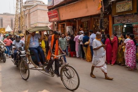 Varanasi, India - March 18, 2024: A cycle rickshaw puller transports a man while a line of people, some in traditional clothing, wait outside shops on a busy main street with motorcycles, pedestrians, and Hindi signage visible.のeditorial素材