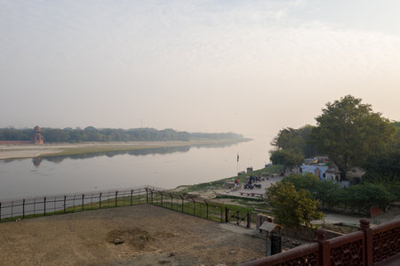 Agra, India - March 11, 2024: The image shows the Yamuna River flowing beside a fenced area with people, motorcycles, and small buildings on the riverbank, with trees and a red sandstone pavilion visible in the distance under a hazy sky.のeditorial素材
