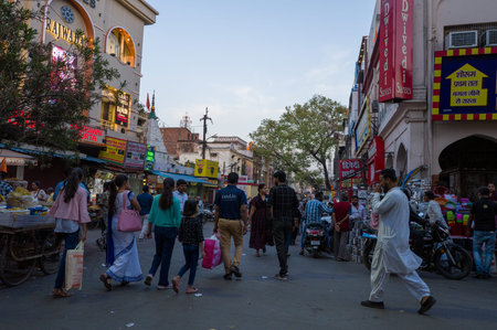 Lucknow, India - March 16, 2024: People walk along a crowded urban street lined with shops, visible signage in Hindi and English, street vendors, and motorcycles during the evening.のeditorial素材