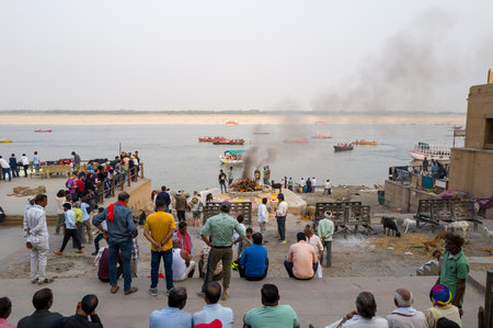 Varanasi, India - March 18, 2024: Groups of people are seated and standing near the steps at Maharaja Harishchandra Ghat, watching cremation rituals taking place along the banks of the Ganges River, with smoke rising from funeral pyres and boats visible oのeditorial素材