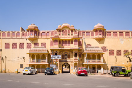 Jaipur, India - March 16, 2022: The main entrance facade of City Palace with traditional Rajasthani architecture, arched windows, domed pavilions, Hindi signage above the entrance, and several parked cars and a jeep on the street under clear daylight.のeditorial素材