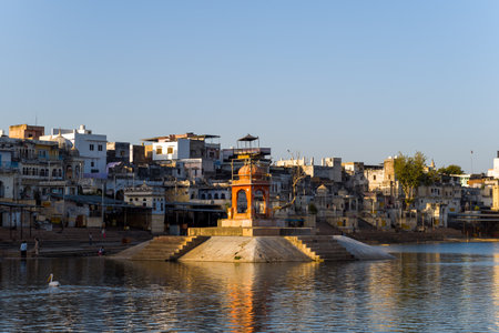 Pushkar, India - March 17, 2022: A central pavilion stands on a platform in the middle of Pushkar Lake, surrounded by water, with stone steps leading to the lake and historic buildings lining the waterfront under clear daylight.のeditorial素材