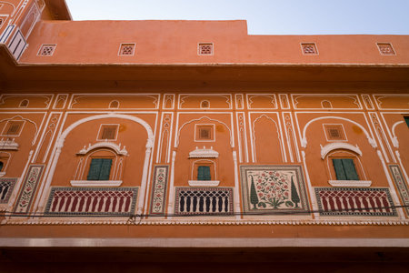 Jaipur, India - March 15, 2022: Detailed view of an orange building facade in the old city featuring traditional arched windows, green shutters, white decorative trim, and painted floral panels under daylight.のeditorial素材