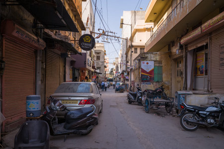 Jaipur, India - March 15, 2022: Narrow street in the old city with closed shop shutters, parked scooters, motorcycles, a car, visible business signage including a coffee shop sign, and pedestrians in the distance under daylight.のeditorial素材
