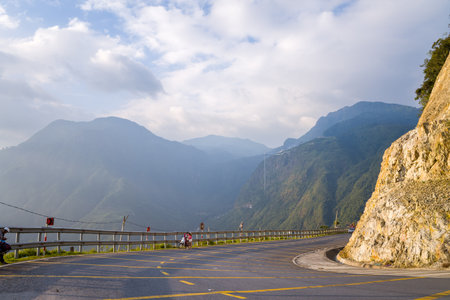Route Lai Chau vers Sapa, Vietnam - September 7, 2022: A curved paved mountain road with yellow markings and a metal guardrail runs alongside a rocky cliff, with two motorbikes and riders stopped at the roadside and misty green mountains in the backgroundのeditorial素材