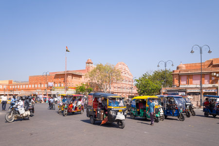 Jaipur, India - March 16, 2022: Multiple auto rickshaws, motorcycles, and pedestrians are seen at a busy intersection with the pink facade of Hawa Mahal and surrounding buildings visible in the background under clear daylight.のeditorial素材