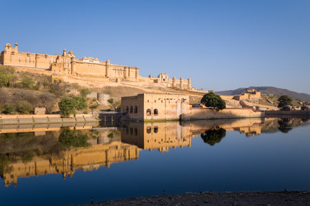 Jaipur, India - March 17, 2022: Amber Fort is visible on a hillside with its sandstone walls and structures reflected in the calm waters of Maota Lake under a clear blue sky.のeditorial素材