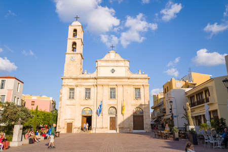 La Canee, Greece - October 21, 2022: The Cathedral of the Virgin Mary, featuring a prominent bell tower and Greek flags, stands in a sunlit square with people sitting and walking nearby, surrounded by residential and commercial buildings under a partly clのeditorial素材