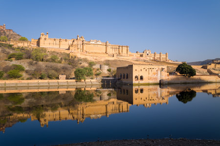 Jaipur, India - March 17, 2022: Amber Fort is visible on a hillside with its sandstone walls and structures reflected in the calm waters of Maota Lake under a clear blue sky.のeditorial素材