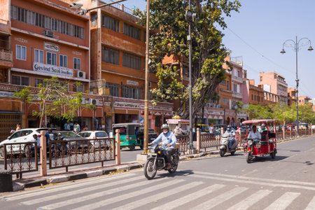 Jaipur, India - March 15, 2022: Motorcyclists, an auto rickshaw, a red electric rickshaw, pedestrians, and parked vehicles are visible on a city street lined with historic pink-painted buildings, Hindi signage, a large tree, and ornate lamp posts under a のeditorial素材