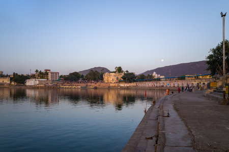 Pushkar, India - March 17, 2022: Pushkar Lake is shown at dusk with several ghats, historic buildings, people gathered along the waterfront, and hills in the background under a clear sky.のeditorial素材