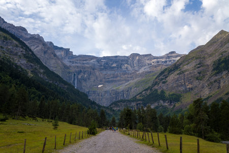 Chemin de randonnee du Cirque de Gavarnie, France - August 9, 2022: A gravel hiking path bordered by wooden posts leads through a grassy valley toward the steep cliffs and waterfall of the Cirque de Gavarnie, with groups of people visible in the distance のeditorial素材
