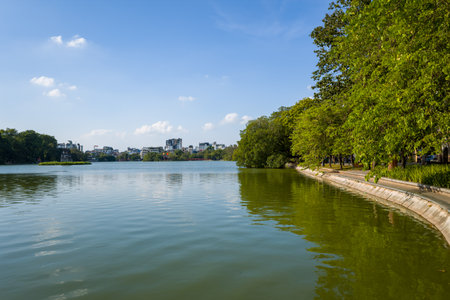 Hanoi, Vietnam - August 29, 2022: The image shows the calm waters of Hoan Kiem Lake with a tree-lined paved walkway on the right and city buildings visible across the lake under a clear blue sky.のeditorial素材
