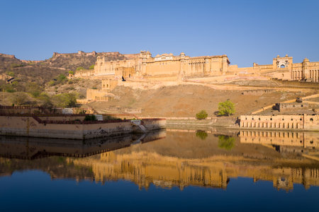 Jaipur, India - March 17, 2022: The Amber Fort is seen on a hillside with its walls and structures reflected in the calm waters of Maota Lake under a clear blue sky.のeditorial素材