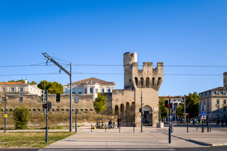 Avignon, France - September 21, 2022: A medieval stone city gate and fortified wall are shown with tram lines, traffic lights, pedestrians, and surrounding buildings under a clear blue sky.のeditorial素材