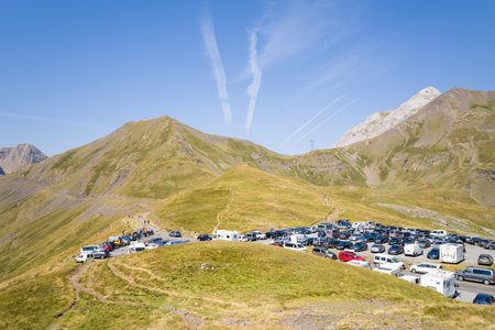 Parking dans les montagnes des Pyrenees, France - August 7, 2022: A parking area with numerous cars, vans, camper vans, and people is situated on a grassy mountain ridge surrounded by high peaks under a clear blue sky with visible contrails.のeditorial素材