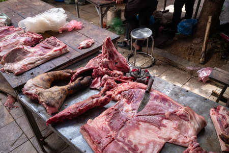 Marche de Bac Ha, Vietnam - September 11, 2022: Butchered pig meat, including a pigs head and leg, is displayed on metal and wooden tables with a large knife at Bac Ha Market, with people and market items visible in the background.のeditorial素材