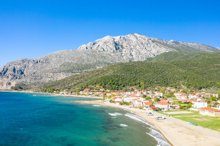 Kato Vasiliki, Greece - October 18, 2022: An aerial view shows the coastal village of Kato Vasiliki with red-roofed houses along the shoreline, a pebble beach, clear blue sea, and a large rocky mountain in the background under a clear sky.のeditorial素材