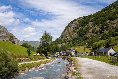 Gavarnie, France - August 9, 2022: A view of a river running alongside a gravel path with rural houses and farm buildings at the base of green mountains under a partly cloudy sky.のeditorial素材