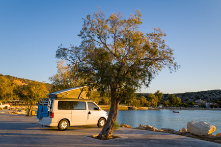 Port de Marathi, Greece - October 23, 2022: A white camper van with an elevated roof is parked beside a tamarisk tree along the paved waterfront at Port de Marathi, with calm water, distant hills, and coastal buildings visible in the background during golのeditorial素材