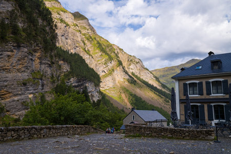 Refuge du Cirque de Gavarnie, France - August 9, 2022: A view of mountain refuge buildings with a stone wall in the foreground, set against steep rocky cliffs and forested slopes under a partly cloudy sky.のeditorial素材