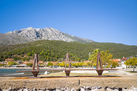 Kato Vasiliki, Greece - October 18, 2022: A metal sculpture representing a cable-stayed bridge is installed on the waterfront promenade with village houses, trees, and a mountainous landscape in the background under a clear blue sky.のeditorial素材