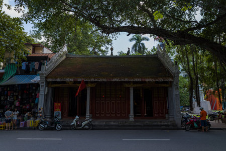 Hanoi, Vietnam - August 29, 2022: The image shows the facade of Den Ba Kieu Temple with a red flag, parked motorbikes, and people standing and sitting nearby, with trees and market stalls visible on the side.のeditorial素材