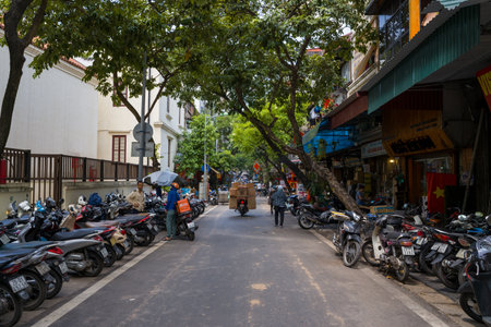 Hanoi, Vietnam - August 29, 2022: The image shows a street lined with parked motorbikes on both sides, a delivery worker with a box and orange backpack, pedestrians, shops with Vietnamese signage, and trees providing shade under daylight.のeditorial素材