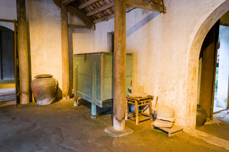 Hanoi, Vietnam - September 15, 2022: The interior of a traditional Vietnamese house is displayed at the Museum of Ethnology, showing wooden pillars, an arched doorway, a large ceramic jar, a green wooden cabinet, and stone grinding tools on stools.のeditorial素材