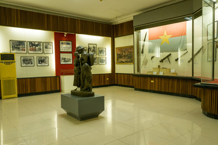 Hanoi, Vietnam - September 13, 2022: Interior view of an exhibition room at the National Museum of History featuring a bronze statue on a pedestal, framed historical photographs on the wall, and a glass display case with rifles, spears, and a large flag.のeditorial素材