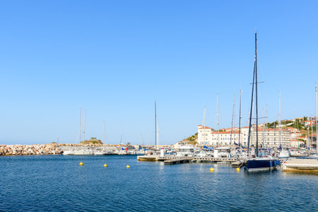 Unknown location - June 19, 2022: The image shows a marina with numerous sailboats and yachts docked at floating piers, with waterfront residential buildings and hillside houses visible in the background under a clear blue sky.のeditorial素材