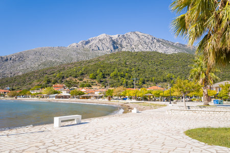 Plage de Kato Vasiliki, Greece - October 18, 2022: A paved promenade with a white bench, palm tree, and street lamps runs along the pebble beach, with village houses and a mountainous landscape in the background under a clear blue sky.のeditorial素材