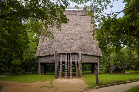 Hanoi, Vietnam - September 15, 2022: The front of a traditional Bahnar communal house is shown with a very tall thatched roof, wooden stilts, and two wooden staircases, surrounded by trees and grass at the Museum of Ethnology.のeditorial素材