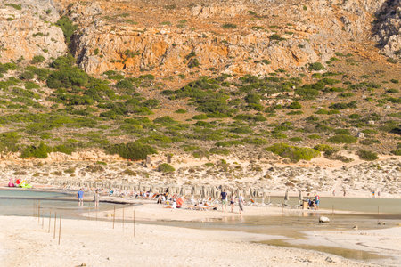 Plage de Balos, Greece - October 24, 2022: Groups of people are visible on the sandy shoreline near sun loungers and closed umbrellas, with a rocky hillside and sparse vegetation in the background.のeditorial素材