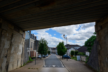 Pouilly sur Loire, France - June 27, 2022: A view from under a stone and concrete railway bridge shows a quiet street lined with residential buildings, parked cars, trees, and a street lamp under a partly cloudy sky.のeditorial素材